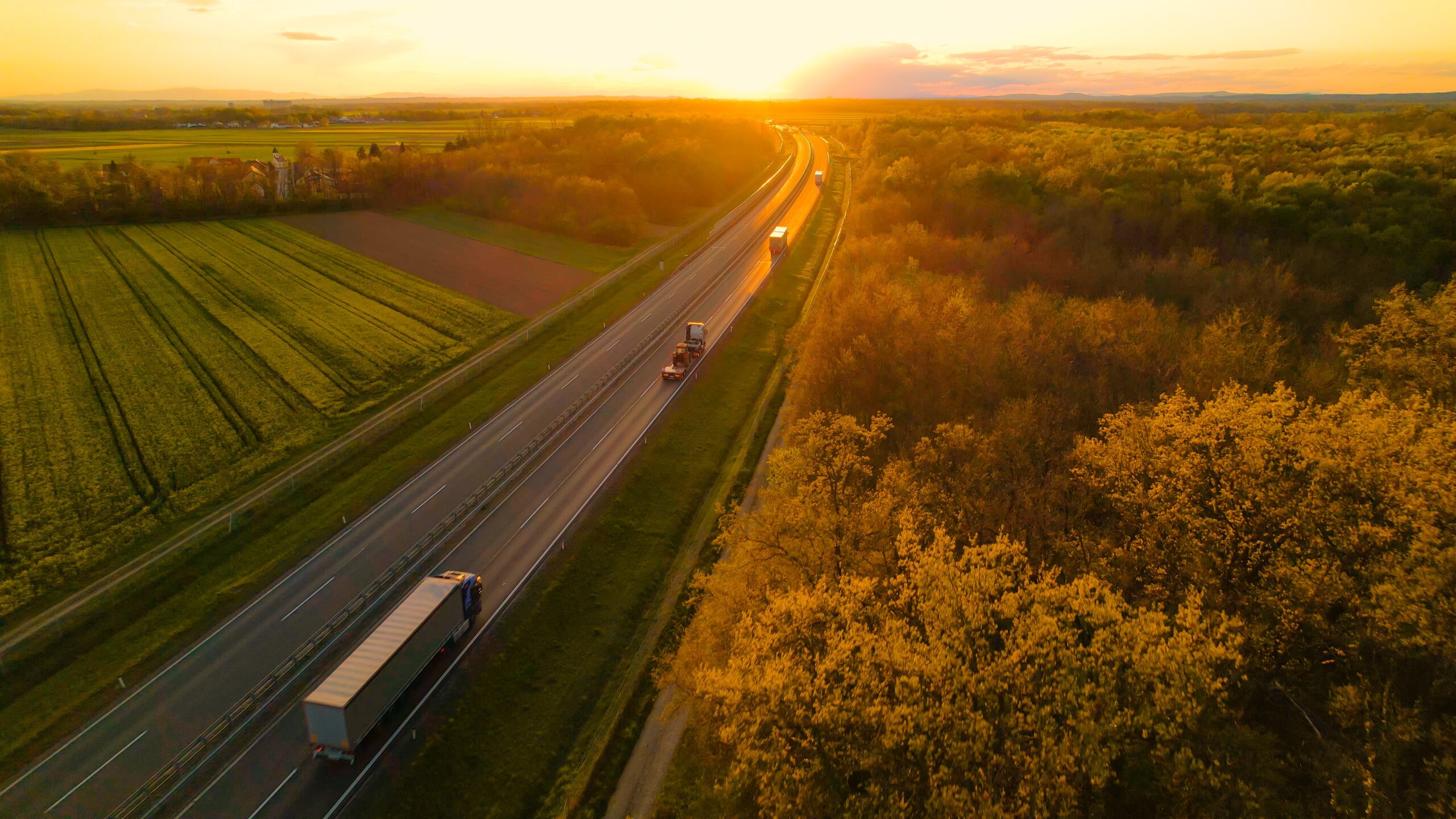 Trucks driving down road near farm fields with sunset in background